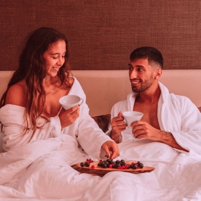 Casal de robe branco na cama com chávenas e tábua de frutas
