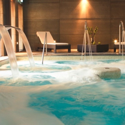 Piscina interior com jatos de água, espreguiçadeiras e decoração numa sala moderna.