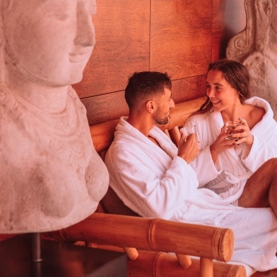 Casal com robes brancos sentados em banco de madeira entre bustos em pedra