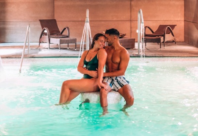 Casal sentado num banco dentro de piscina interior com espreguiçadeiras e torneiras de água ao fundo