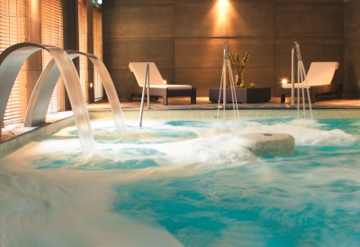 Piscina interior com jatos de água, espreguiçadeiras e decoração numa sala moderna.