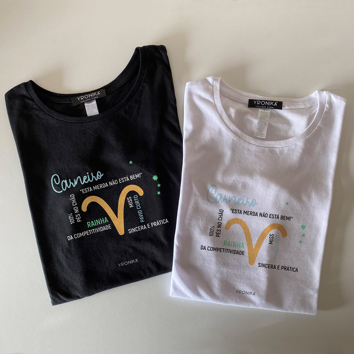 Signo Carneiro Duas t-shirts, preta e branca, com estampas coloridas e texto, em superfície clara.