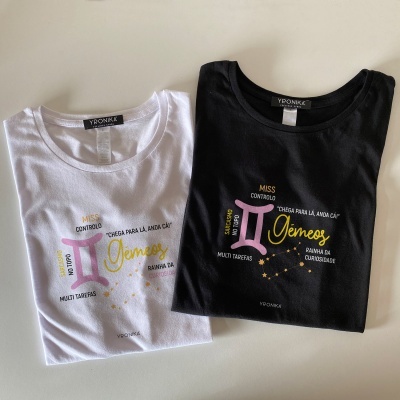 Duas t-shirts, uma branca e outra preta, com estampas coloridas do signo Gêmeos e texto em português