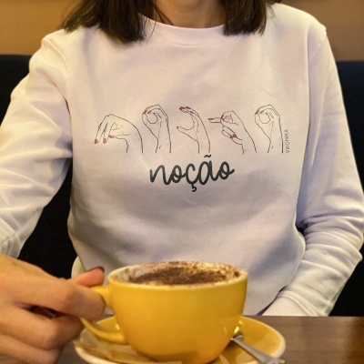 Camisola branca com estampa de mãos e palavra 'noção' com pessoa e chávena amarela de café