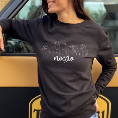 Mulher com sweatshirt preta com ilustrações de mãos e palavra 'noção' em branco junto a carro preto e bege