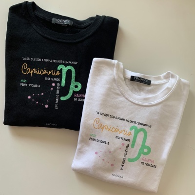 Duas t-shirts dobradas, preta e branca, com estampas do signo Capricórnio e texto em português.