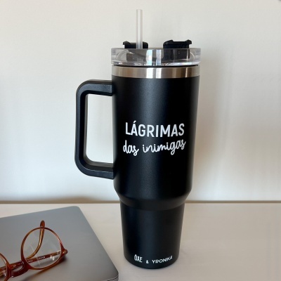 Copo térmico preto com texto em branco e tampa com canudo, ao lado de um portátil e óculos.