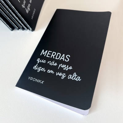 Caderno preto com texto branco 'MERDAS que não posso dizer em voz alta' e assinatura VPRONIKA