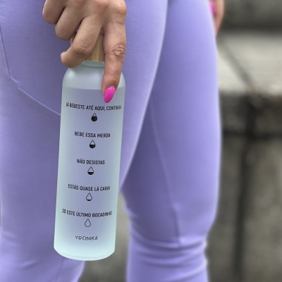 Garrafa de água transparente com texto em português e tampa de madeira sendo segurada em frente a calças lilás