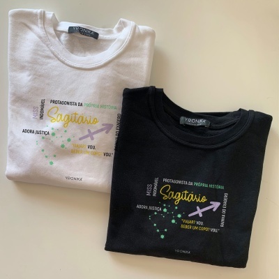 Duas t-shirts dobradas, branca e preta, com texto e símbolos do signo Sagitário.