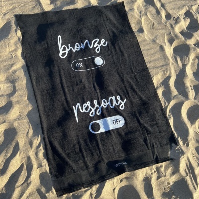 Toalha preta com texto humorístico branco na areia