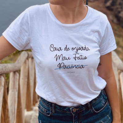 Pessoa de camisa branca com texto em português posando num ambiente exterior com corrimão de madeira.