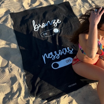 Toalha preta com texto 'bronze ON' e 'pessoas OFF' em areia de praia junto a pessoa em biquíni colorido