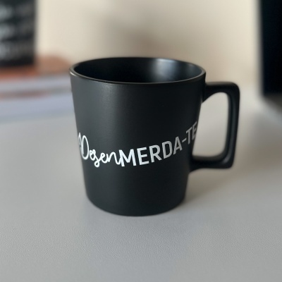 Caneca preta com inscrição branca OsenMERDA-TE sobre superfície cinza claro