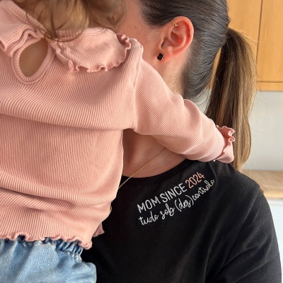Camisola preta com bordado MOM SINCE 2024 usada por mulher com criança abraçada em camisa rosa e ganga azul