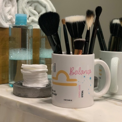 Caneca com pincéis de maquilhagem numa bancada de casa de banho com discos de algodão, garrafa de líquido e toalhas