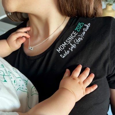T-shirt preta com mensagem bordada MOM SINCE 2025 usada por mulher com criança