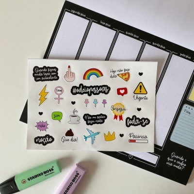 Autocolantes coloridos com texto em português sobre bloco de notas preto e marcadores pastel