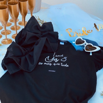 Conjunto despedida de solteira com t-shirt preta, óculos coração, copos dourados e faixa azul 'Bride to'