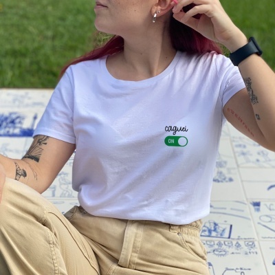 Pessoa sentada com t-shirt branca com frase 'caquei ON' e calças bege, sobre azulejos decorativos no chão
