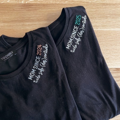 Duas t-shirts pretas bordadas com textos em várias cores, sobre madeira clara