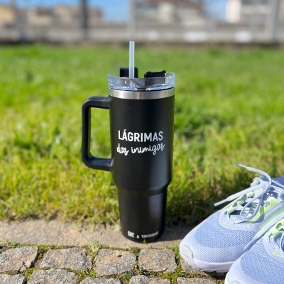 Copo térmico preto com pega e tampa transparente e ténis azuis sobre calçada e relvado