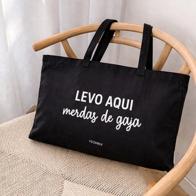 Saco de pano preto com texto branco colocado numa cadeira de madeira clara.