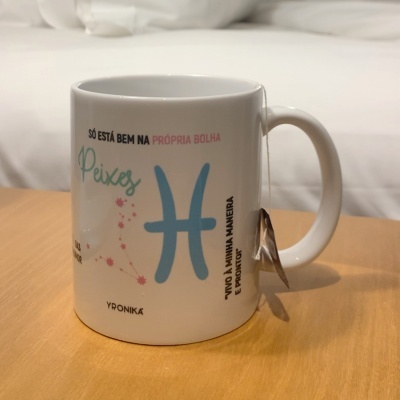 Caneca branca com texto colorido e colher presa à pega sobre mesa de madeira