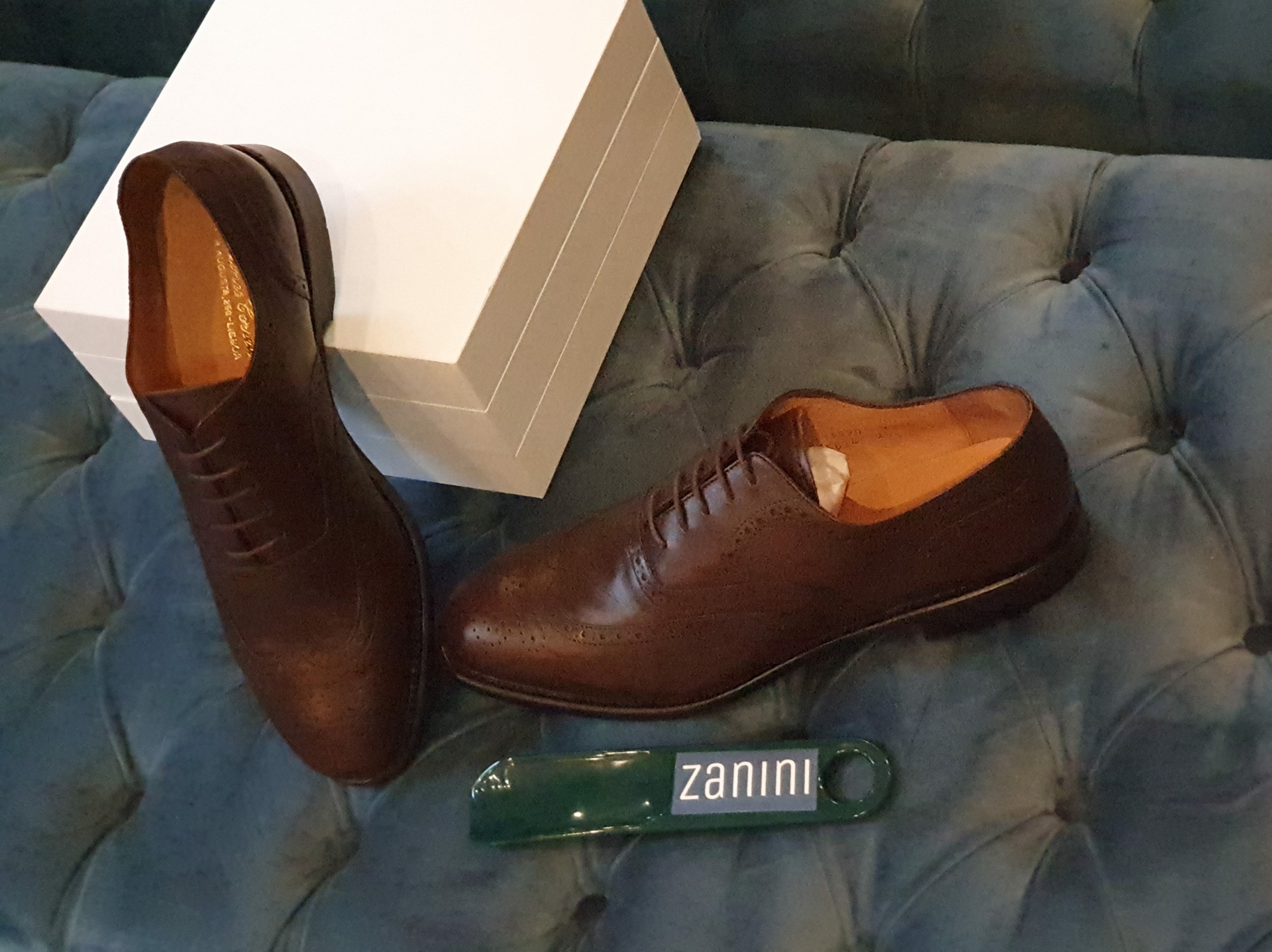 OXFORD WING TIP CASTANHO Sapatos castanhos de pele com cadarço num sofá azul, com caixa branca e sapateiro zanini.