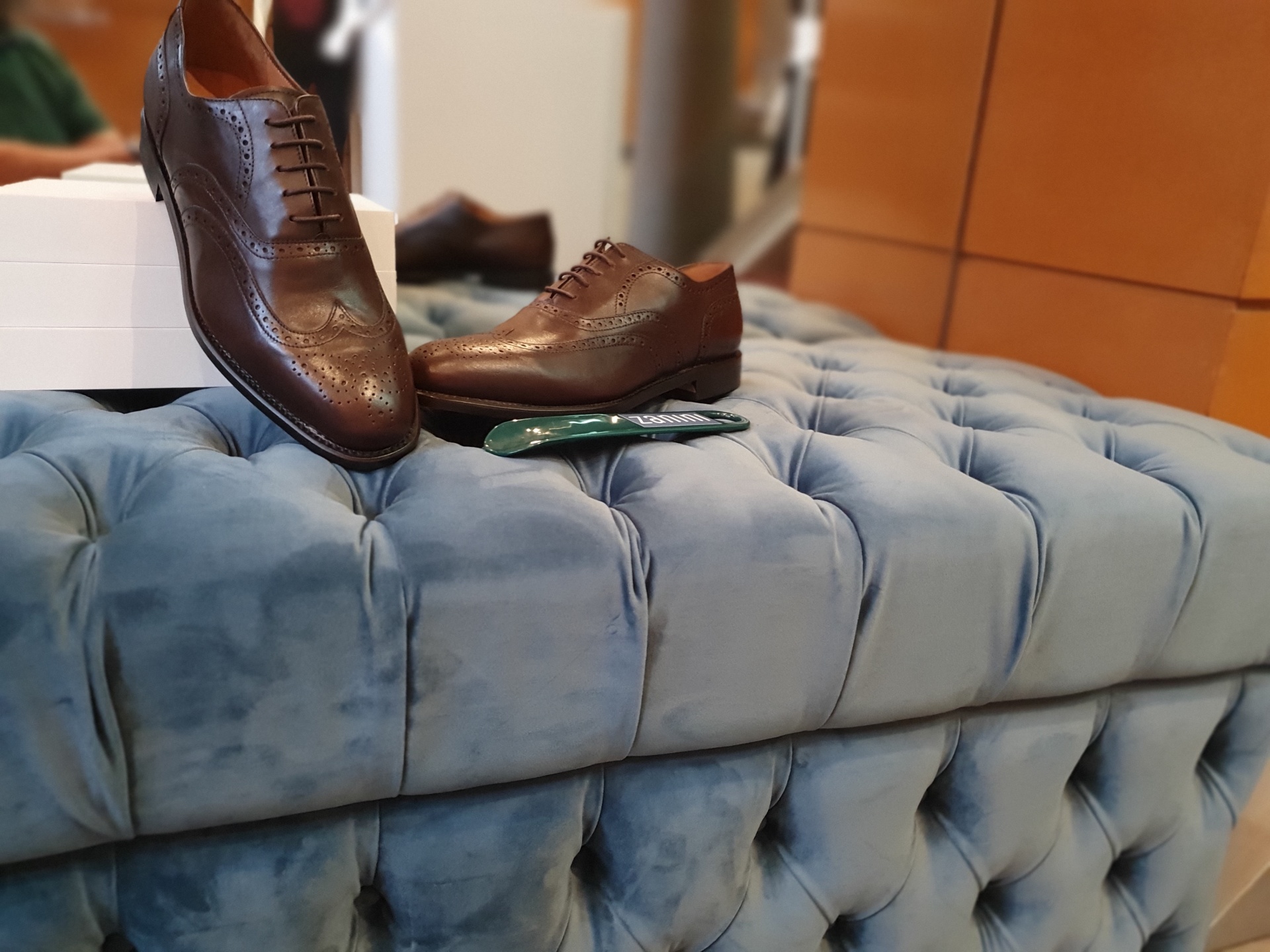 OXFORD WING TIP CASTANHO Sapatos sociais de couro castanho sobre sofá azul claro em loja