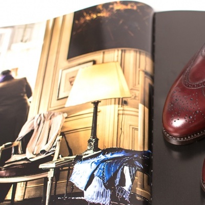 Sapatos de couro castanho com padrão perfurado ao lado de revista aberta com fotografia de interior.