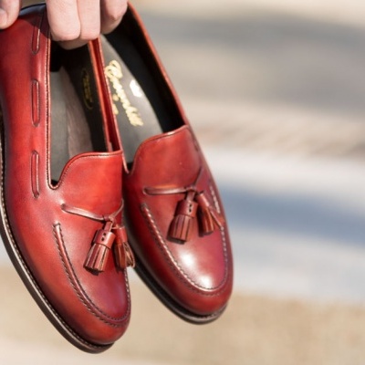 Par de mocassins vermelhos em pele com borlas e costuras, segurados por uma pessoa com calças de ganga azuis.