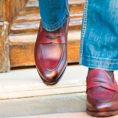 Sapatos masculinos de couro castanho-avermelhado com calças de ganga azuis junto a porta de madeira.