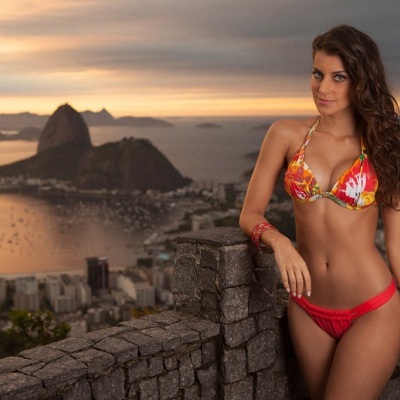 Mulher a usar biquíni vermelho com padrão floral junto a muro de pedra com vista para baía ao pôr do sol