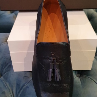 Sapato preto de couro com tassel sobre caixas brancas e logo CENTENÁRIO no fundo