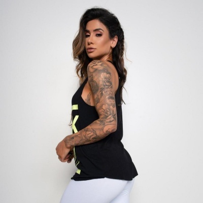 Mulher de lado usando top preto com letras amarelas e calças brancas, tatuagens visíveis no braço.