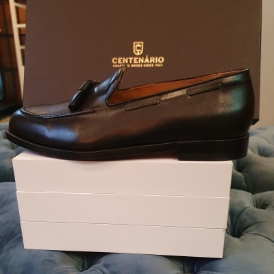 Sapato preto tipo mocassim em pele sobre caixa branca e caixa castanha com texto CENTENÁRIO