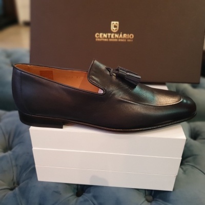 Sapato preto em pele com tassel, em cima de caixa branca, caixa castanha ao fundo com texto CENTENÁRIO