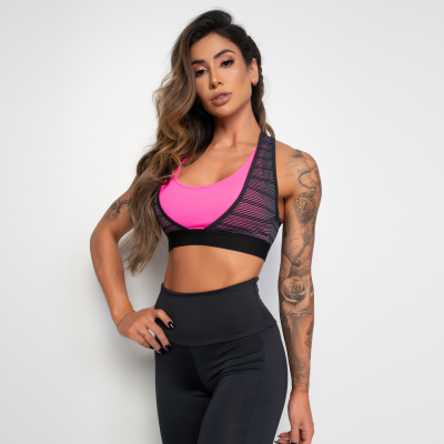 Mulher com top rosa e preto e calças pretas, com tatuagens no braço direito, de pé contra fundo branco.