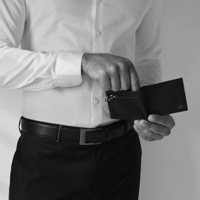 Homem com camisa branca, calças pretas e carteira preta com fecho e iniciais JS