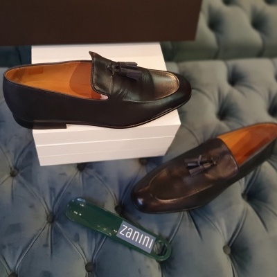 Sapatos de pele preta com tassel sobre caixas brancas e escova verde Zannini numa superfície azul estofada.