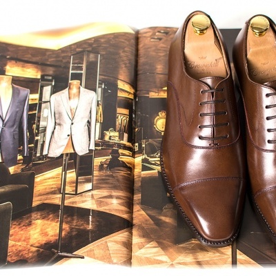 Sapatos sociais castanhos em couro polido sobre revista aberta com fatos masculinos.