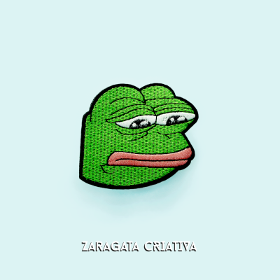 Bordado de cabeça de sapo verde com expressão triste em fundo azul claro e texto ZARAGATA CRIATIVA