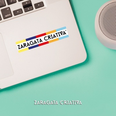 Adesivo colorido 'ZARAGATA CRIATIVA' em portátil prateado numa superfície verde água