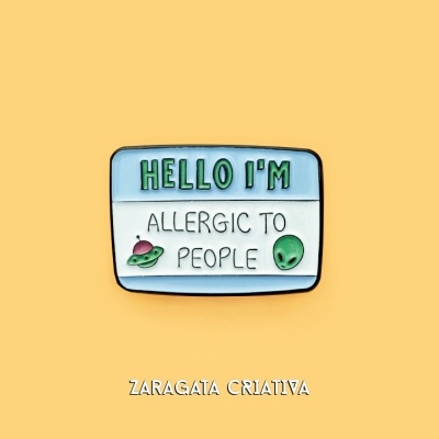 Pin metálico com texto e ícones alienígenas sobre fundo amarelo