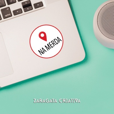 Autocolante redondo com texto 'NA MERDA' num portátil prata sobre fundo azul esverdeado com coluna sonora cinza.