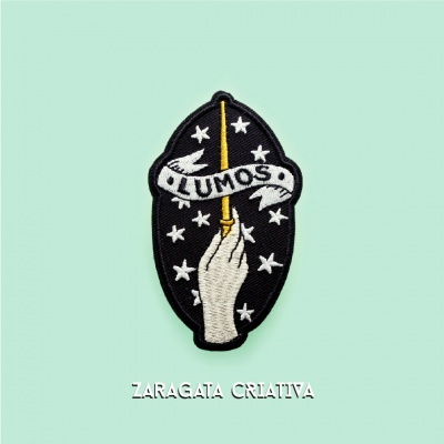 Emblema bordado com mão, varinha mágica amarilla, estrelas brancas e texto LUMOS