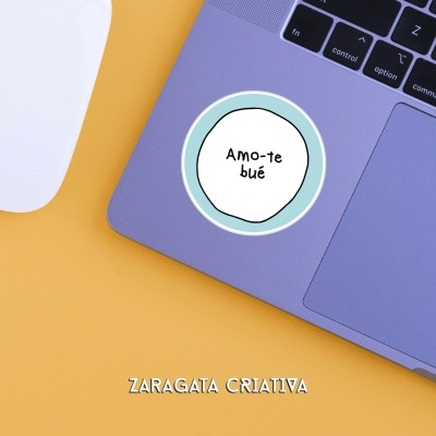 Portátil cinzento-azulado com autocolante 'Amo-te bué' e rato branco sobre superfície amarela.