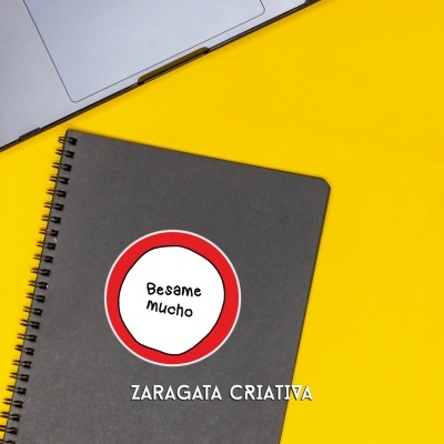 Caderno cinza com autocolante 'Besame Mucho' ao lado de portátil sobre fundo amarelo.