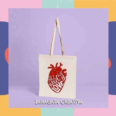 Saco de tecido creme com impressão vermelha em forma de coração e texto, sobre fundo lilás com moldura colorida